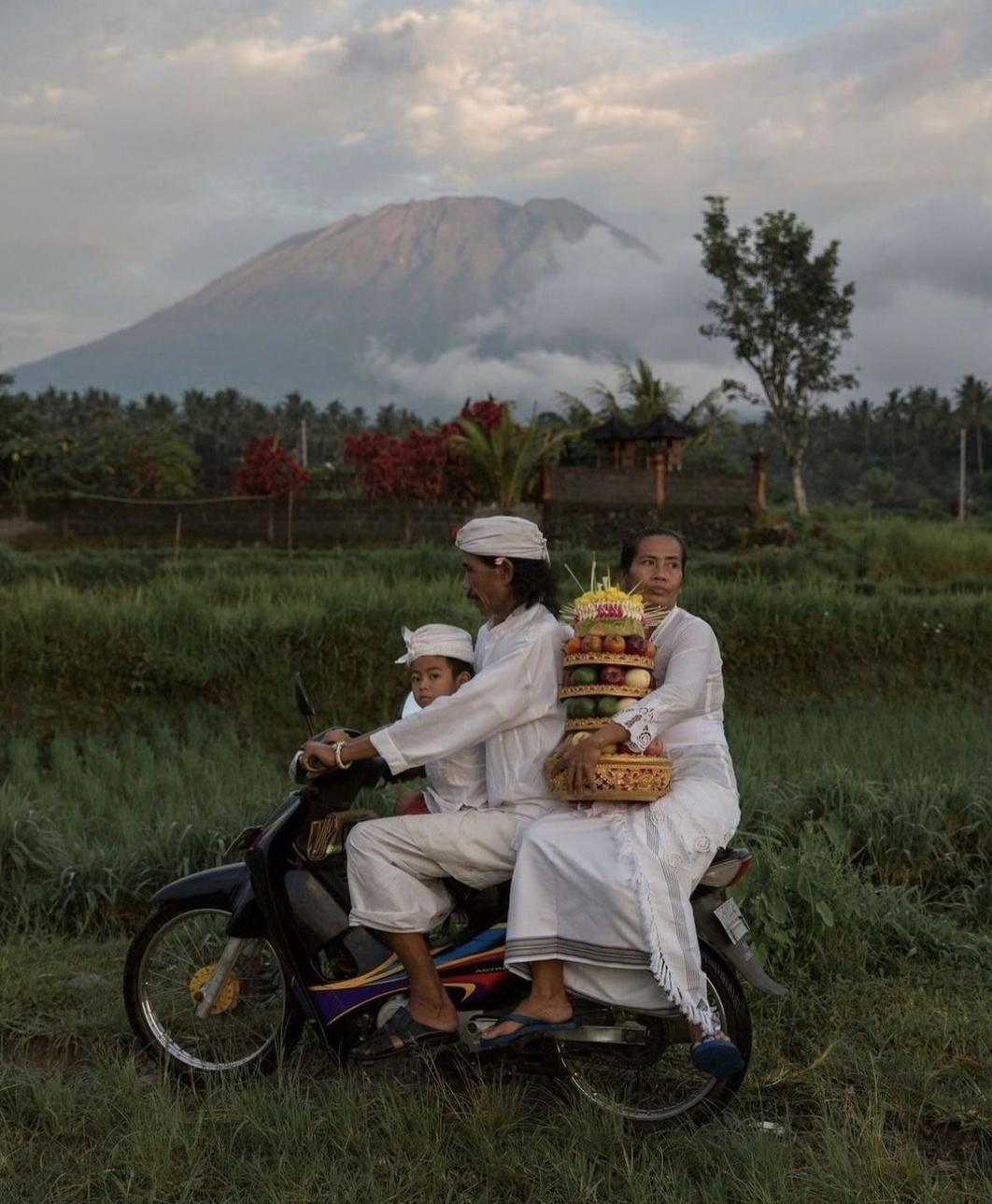 Family in white ceremony dress riding a scooter past rice fields with an offering tower, Mount Agung behind